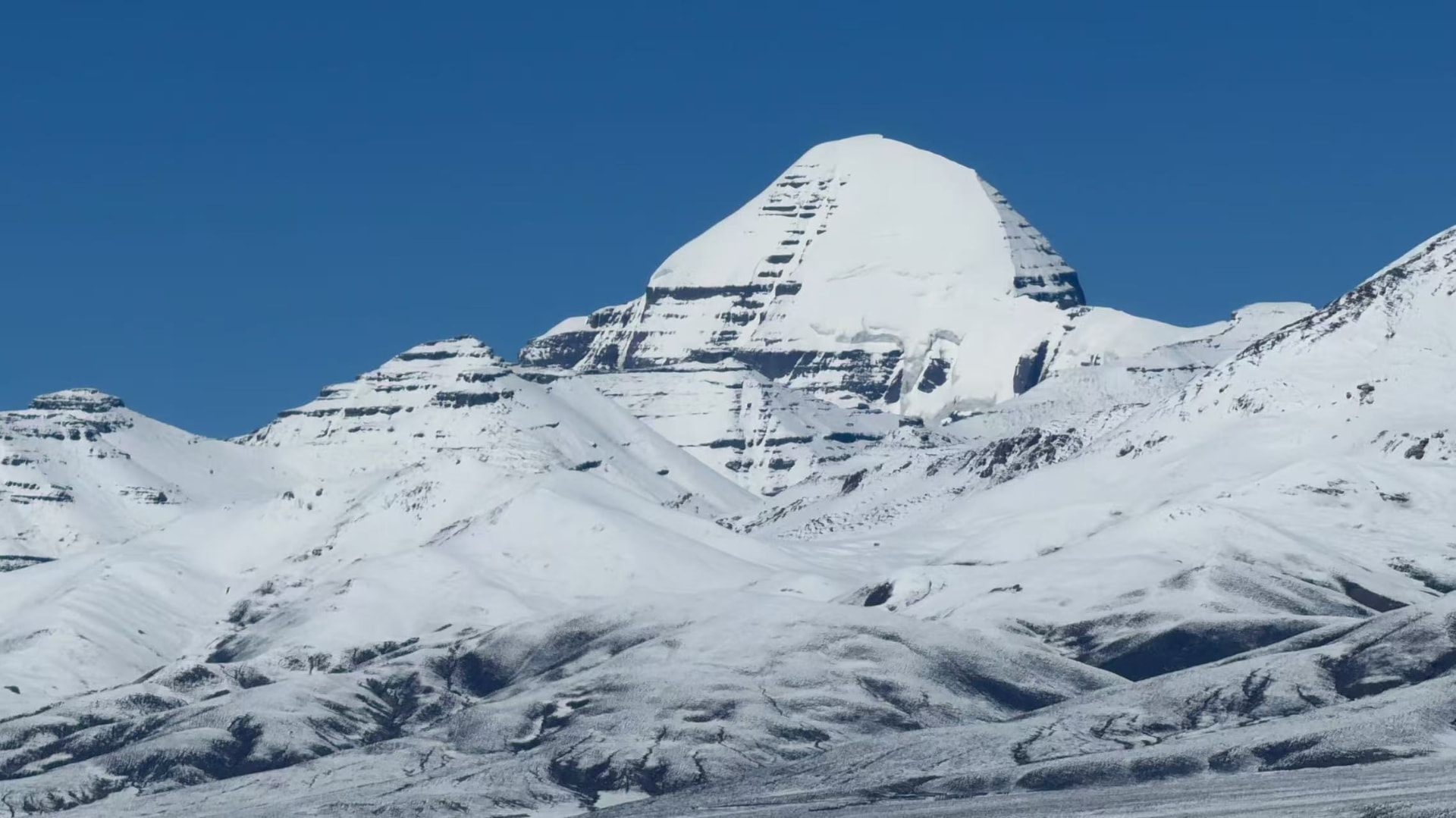 A wide panoramic view of snow-covered Mount Kailash under a clear blue sky