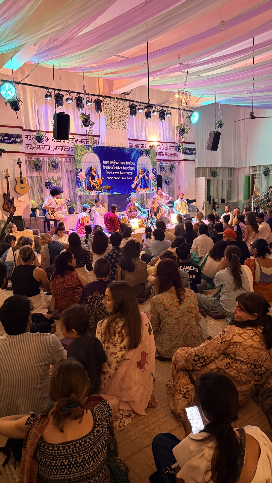 Evening kirtan in Rishikesh, with musicians on stage chanting the Maha Mantra under pastel drapes as devotees sit together on the floor.